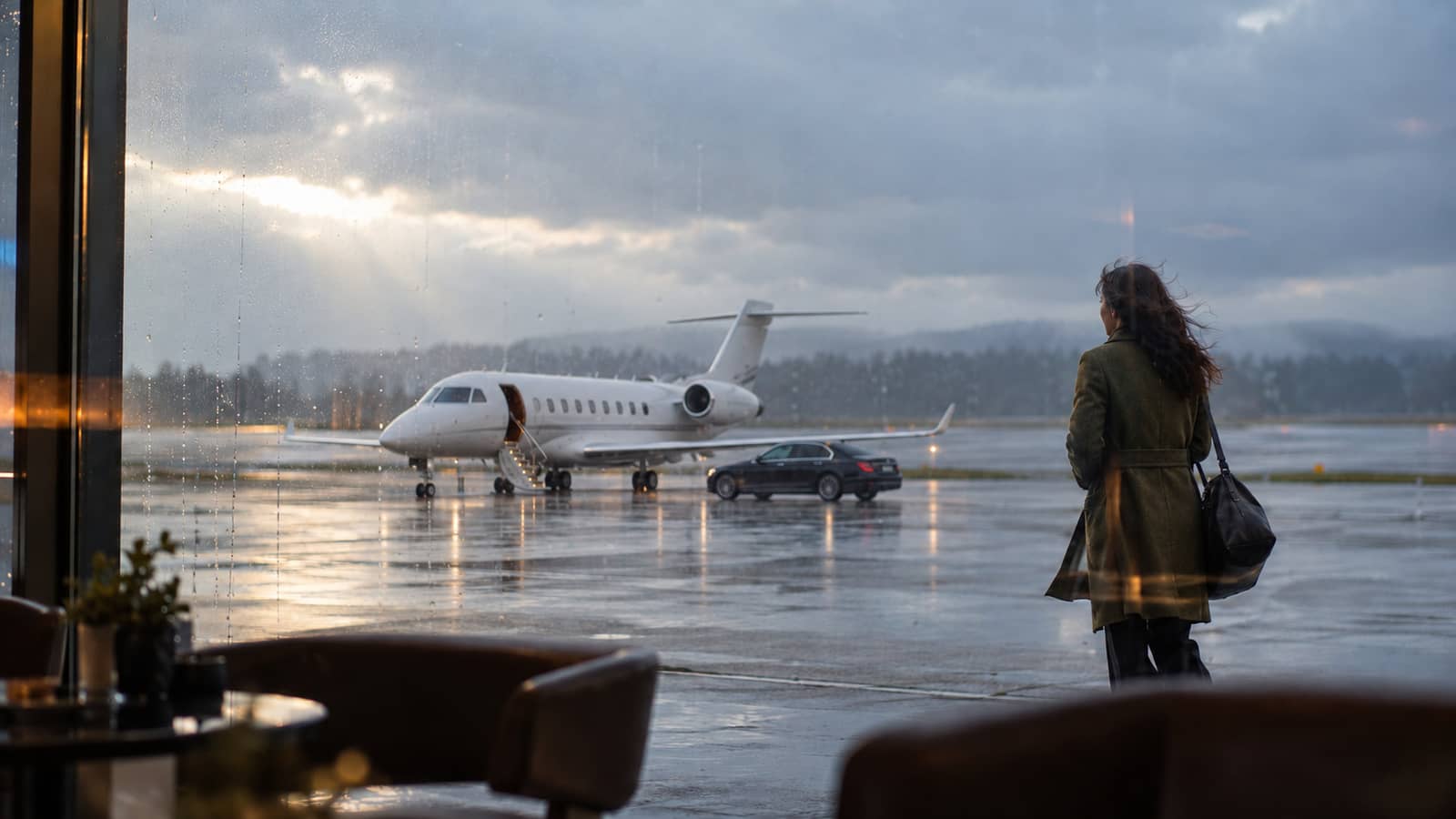 View from a dim terminal lounge of a business jet and a black car on a rainy apron