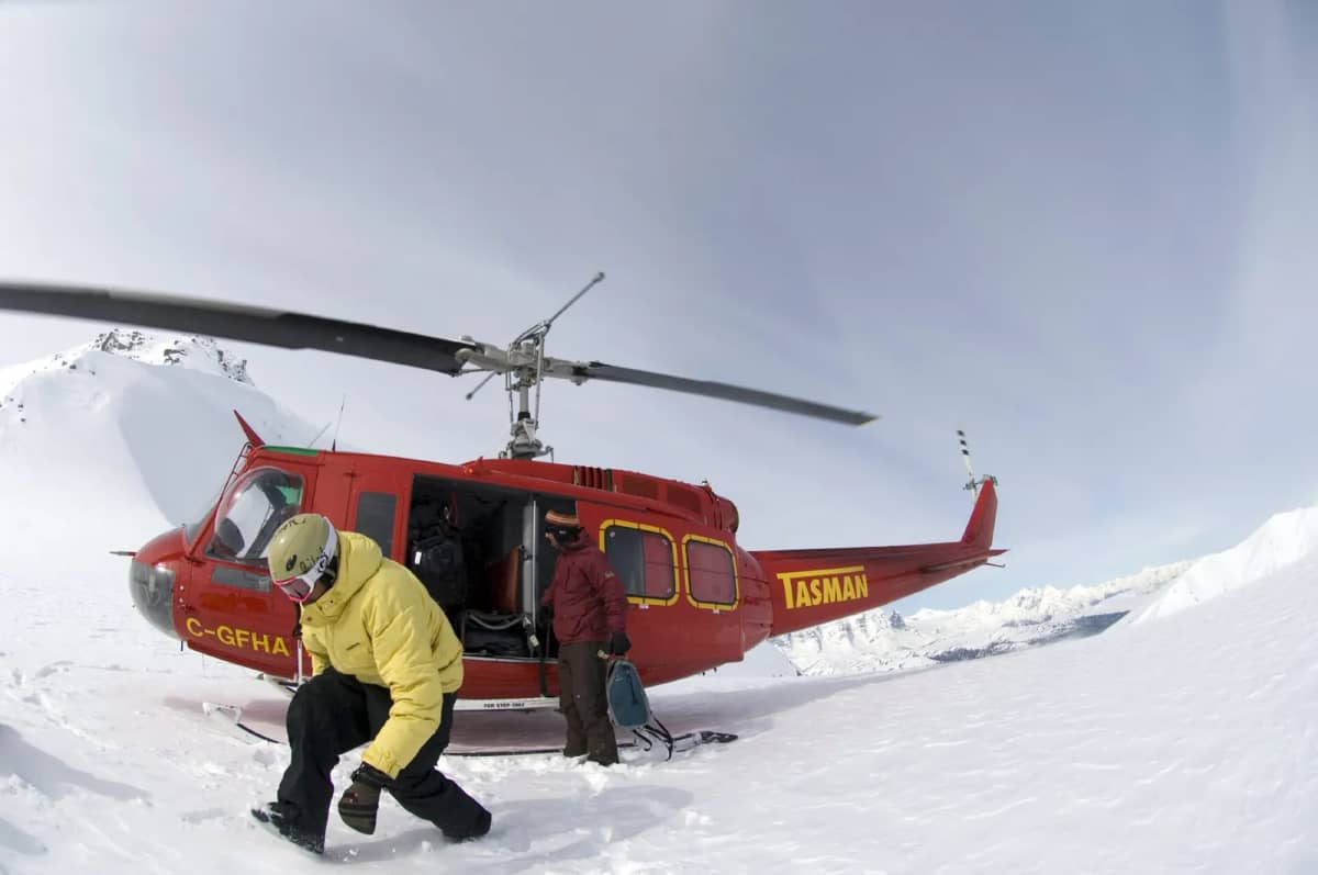 A red Bell 205 helicopter on a snowy peak while skiers step out